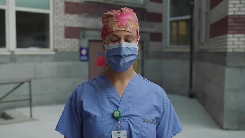 A woman in scrubs mask and cap wearing an id stands outside of a hospital