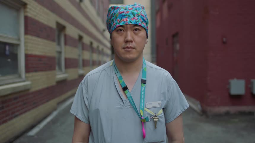 A nurse in scrubs stands in an alley next to brick buildings