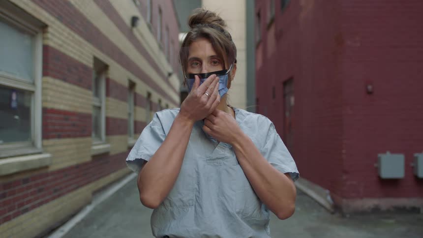 A nurse puts on a surgical mask while standing outside in an alley