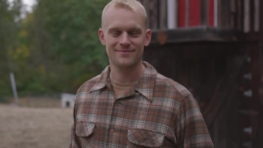 A young man in a flannel shirt stands outside near a building