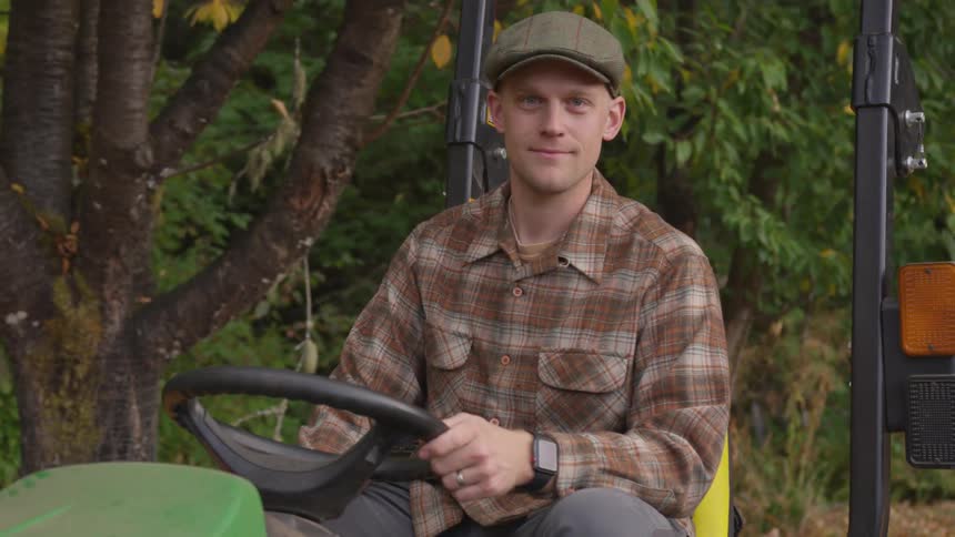 Man sits on riding lawnmower near trees smiling slightly