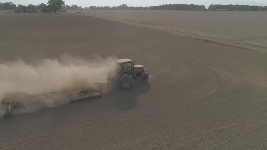 Tractor tows plow across empty crop field kicking up a cloud of dust