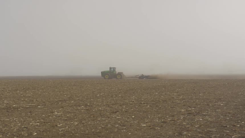 A person drives a green tractor pulling a trailer through a farm field