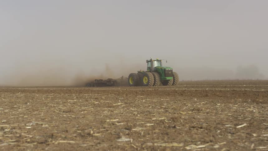 Tractor tows plow across bare field kicking up dust