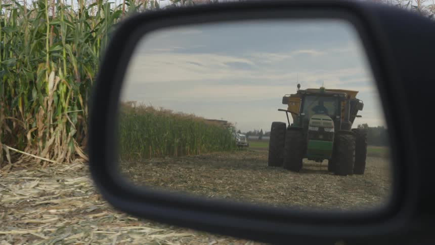 Truck loaded with corn on a farm field reflected in the rearview mirror of a car