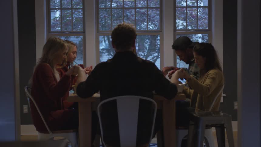 Hosts and dinner guests hold hands and pray at dining table near window showing snow