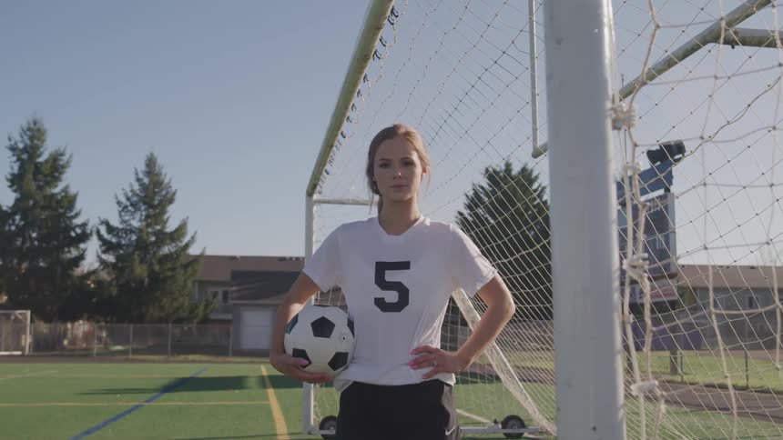 A young female athlete looks into the camera while holding a soccer ball and standing in front of a soccer goal on an athletics field