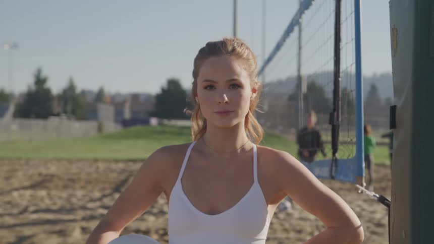 A teenager stands on a sand volleyball court holding a ball as others wait on the court