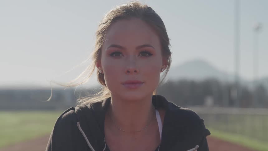 A young woman standing on an athletics track raises her head to look into the camera