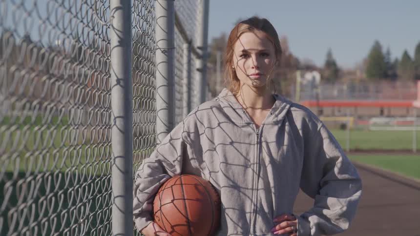 A woman holds a basketball standing by a fence and outdoor track and field