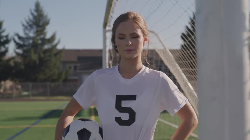 Young woman in sports uniform stands under a goal holding a ball in a soccer field