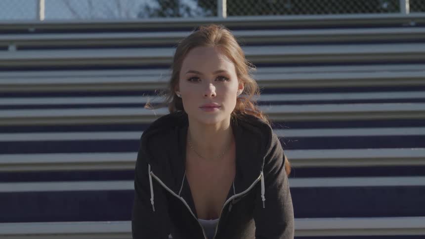 A woman wearing a hoodie sits in the bleachers at an open air stadium