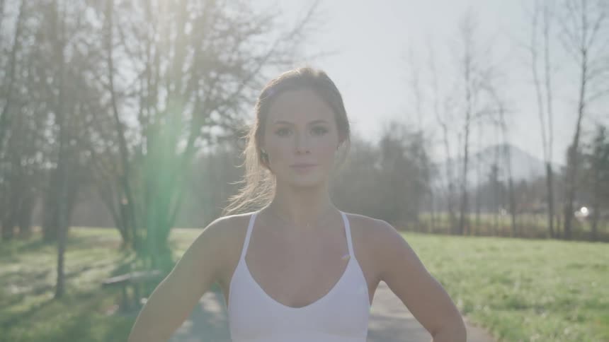A white woman with brown hair is standing in an open field wearing a white tank top