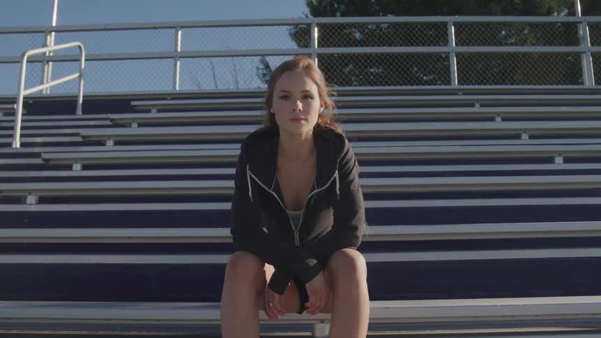 A woman wearing shorts and a black sweatshirt sits on bleachers alone in an empty outdoor stadium