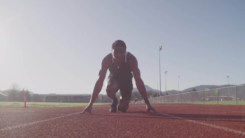 A young black male braces himself in position to begin sprinting on a running track