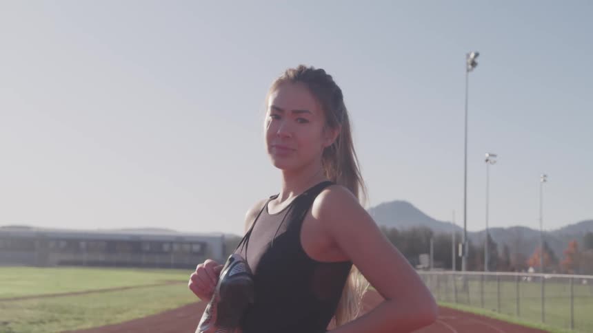 A teenage girl stands on a running track and poses with her running shoes around her neck