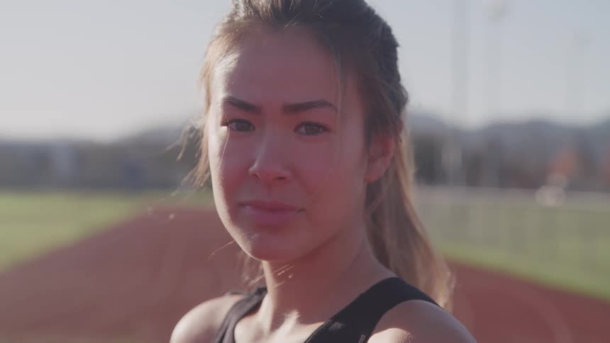 A young woman standing outdoors on an athletics track turns her head to look into the camera