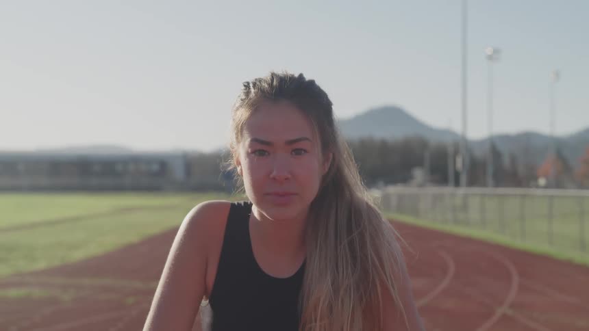 A young woman raises her head to look into the camera while kneeling in preparation for a sprint on an athletics track
