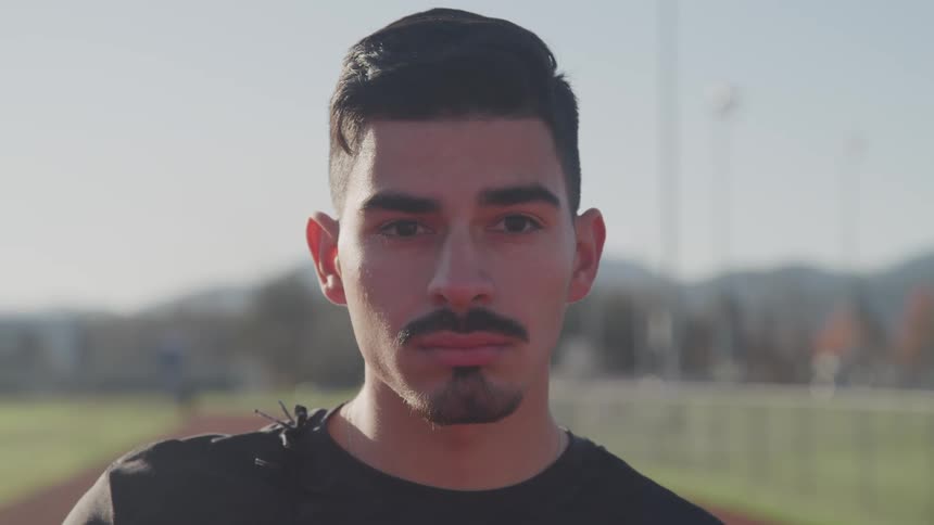 A hispanic male with black hair and a mustache and beard wearing a black shirt is standing on a track