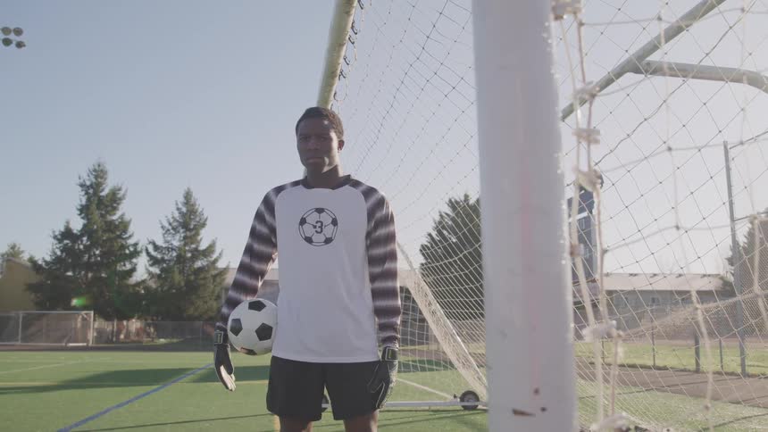 Soccer goalie stands under the goal net with a ball on his arm at morning