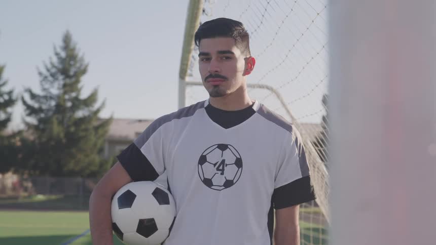 A teenage boy holds a soccer ball as he stands in a soccer net