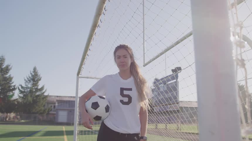 A female stands beside a goal holding a soccer ball