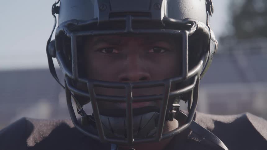 A black male football player wearing a black helmet and jersey is standing on a field