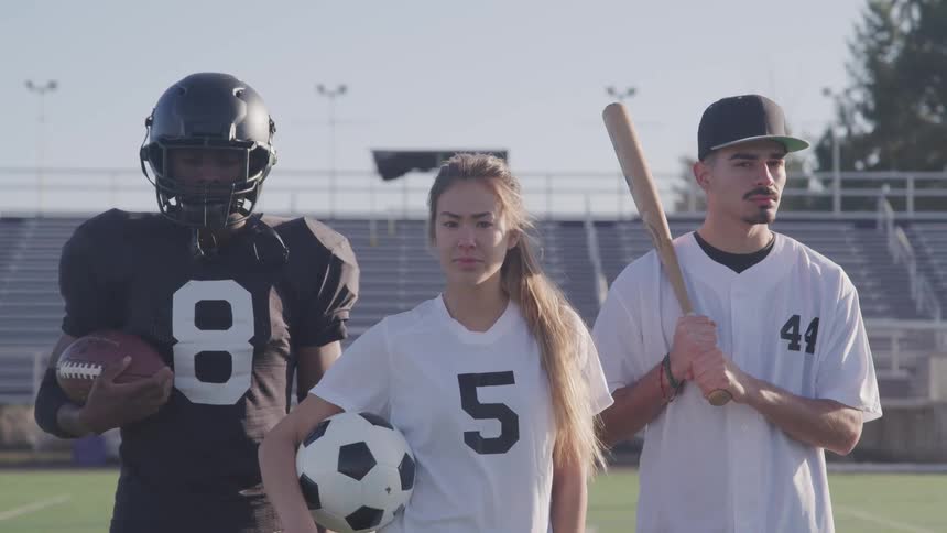 Three sports athlete stand in sports field backed by stadium seating holding sports equipment and wearing jerseys