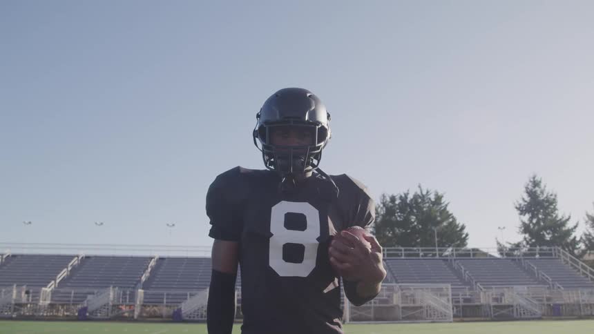 A black man in football gear and holding a football turns his head to look into the camera while standing in front of empty stands on a football field