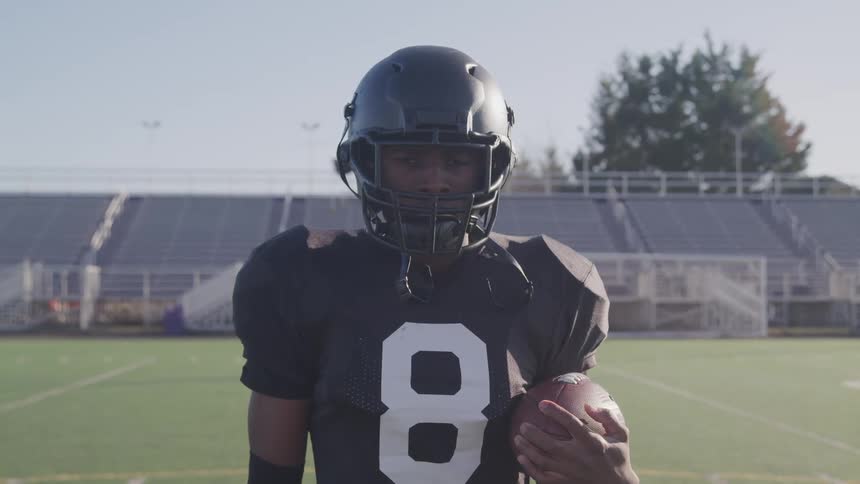Young black man in football jersey and equipment holding football looks to camera