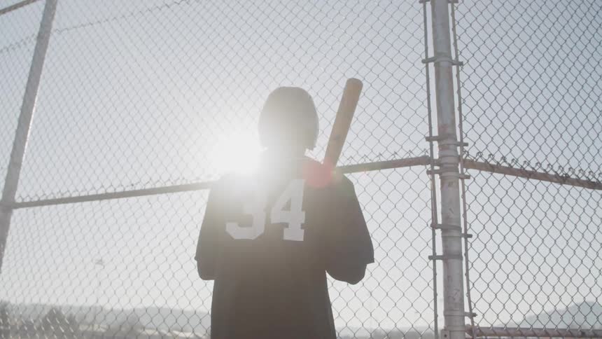 Young man wearing baseball equipment standing near a fence at sunset