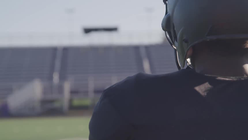Man wearing football helmet and uniform stands in front of a football field in a stadium at day
