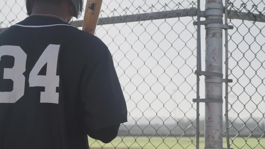 A man holding a baseball bat looks through a fence at a field