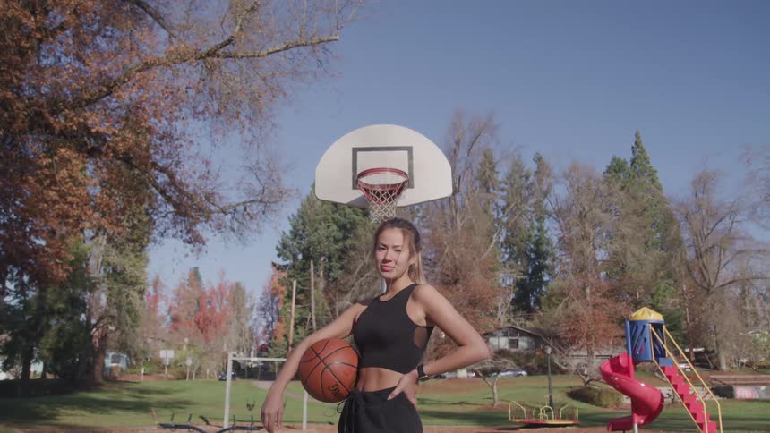 A woman with a basketball on a basketball court with trees and the sky in the background