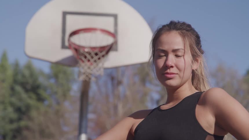 A woman standing and staring on a basketball court with trees and the sky in the background