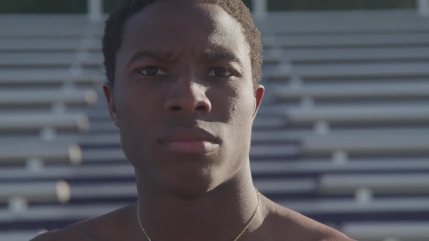 A shirtless young black man standing in front of a set of outdoor bleachers turns his head to gaze into the camera