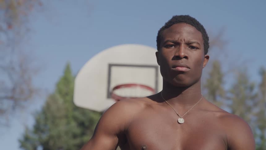 An athletic young black male poses in front of an outdoor basketball hoop