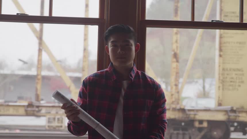 Young man holds blueprint with train visible outside window in background