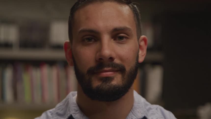 Man with a beard standing in front of a bookshelf faces the camera with eye contact