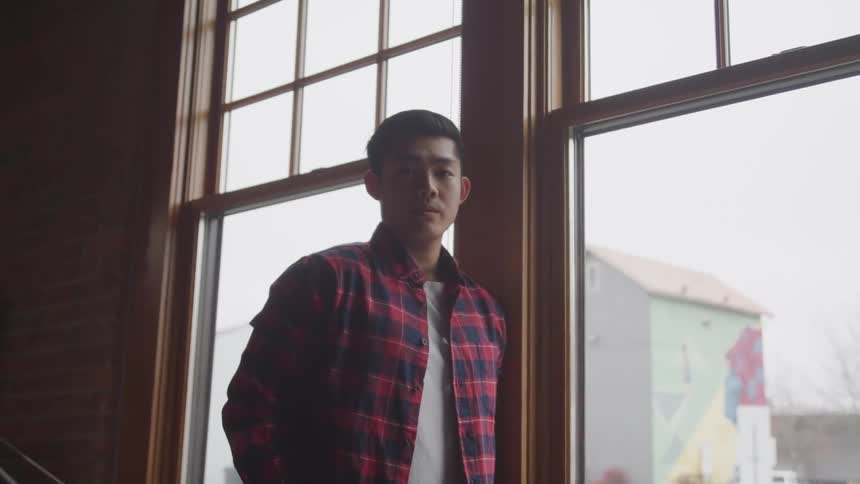 A young asian man stands near a large window while posing for the camera