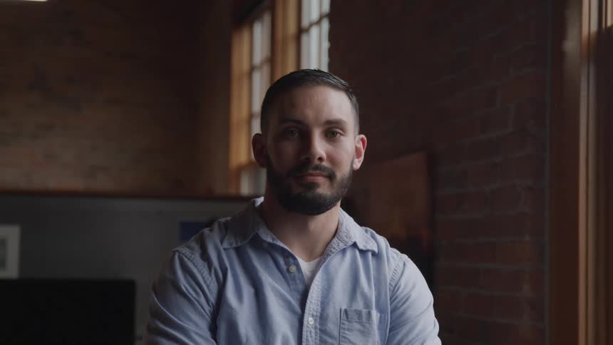 A bearded man in a blue shirt poses for the camera while indoors