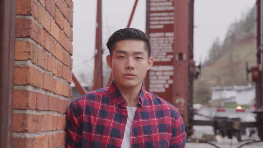Asian man with a plaid shirt leans against a brick wall stares at the camera in the train station