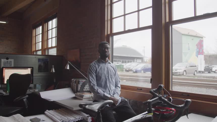 A man sits on the edge of his desk in his office next to a window with views of a train track
