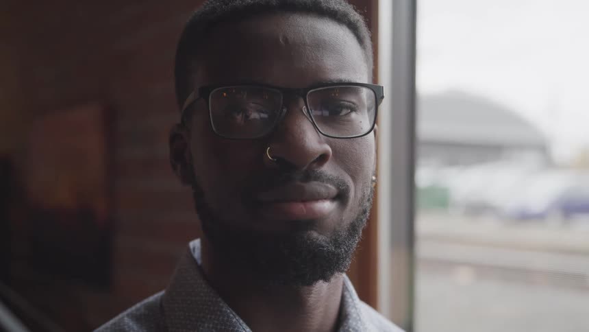 Black man with glasses a nose piercing and a beard looks at the camera smilingly
