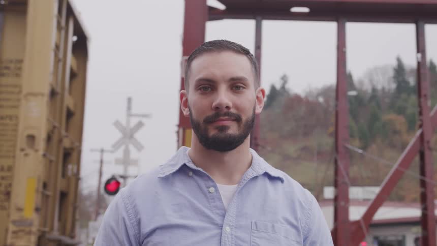 A hispanic male with black hair and black facial hair wearing a blue shirt is standing in front of a train track with a train moving behind him