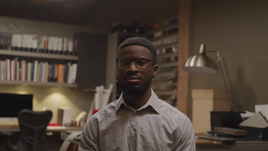 Black man with a shirt a beard and glasses sits down and looks at the camera in a room