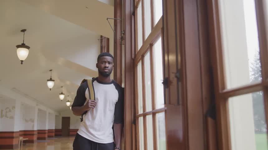 Black man with a white sweatshirt and a backpack leans against windows and faces the camera with eye contact in the corridor