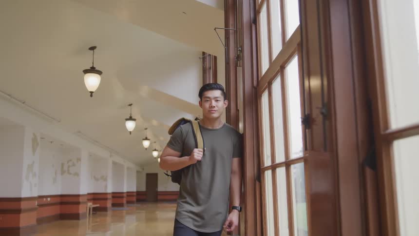 A male college students leans against the large window in the building hallway