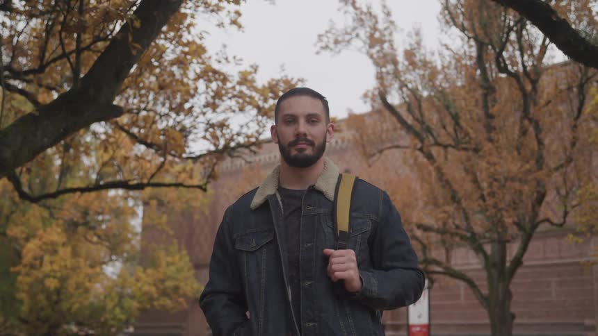 A man with a backpack and a jacket standing with trees and a building in the background