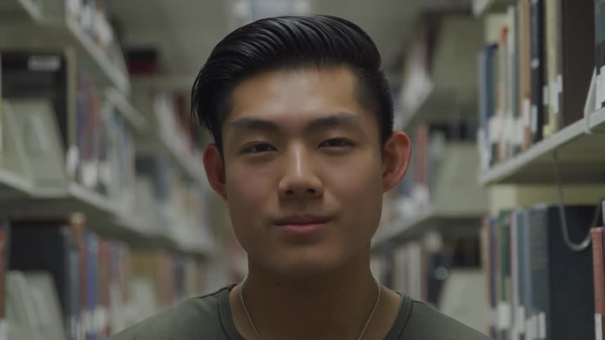A young asian man smiles subtly as he stands inside of a library